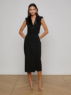 A woman with long brown hair stands indoors against a white wall, wearing the L'AGENCE Hollis Vest—a sleeveless black, power-suit-inspired dress with a deep v-neck and midi-length skirt—paired with earrings and clear high-heeled sandals.