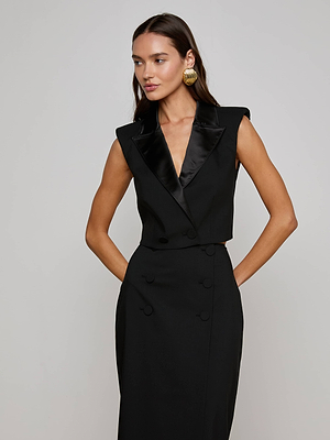 A woman models the Hollis Vest by L'AGENCE— a sleeveless black satin lapel vest— with large gold earrings, standing against a plain light background with her hands in her pockets for a modern power suit look.