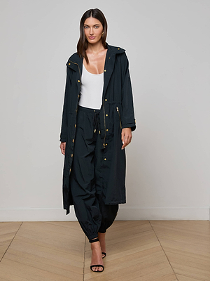 A woman stands indoors in the Toshi Windbreaker, featuring a trench silhouette and gold buttons, layered over a white top with matching black jogger pants and black heeled sandals, set against a plain light background.
