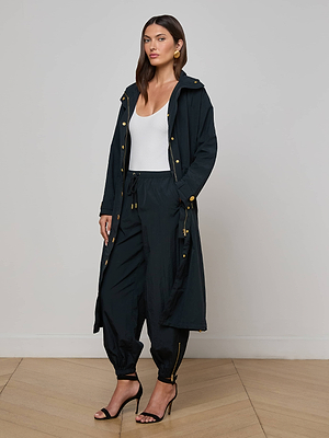 A woman with straight brown hair and gold earrings stands indoors against a plain wall, wearing the Toshi Windbreaker—a sleek long black coat—paired with black jogger pants, a white tank top, and black high-heeled sandals.
