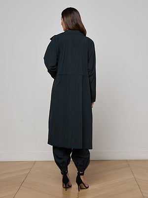 A woman with long brown hair stands facing away, wearing the Toshi Windbreaker and cuffed black pants with black high-heeled sandals, in a minimal indoor setting with light wood flooring.