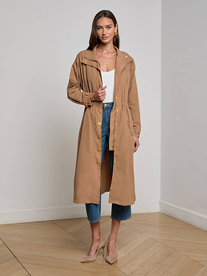 A woman poses indoors in the L'AGENCE Toshi Windbreaker, a long water-repellent trench coat, styled over a white top and cropped blue jeans with beige heels. She stands in a minimalist room with light walls and wood flooring.