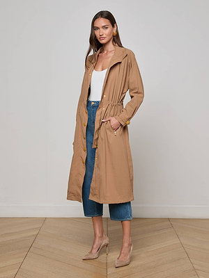 A woman poses confidently indoors wearing the L'AGENCE Toshi Windbreaker, a tan water-repellant trench coat, over a white top and blue cropped jeans. She completes her look with beige heels, gold earrings, and long brown hair.