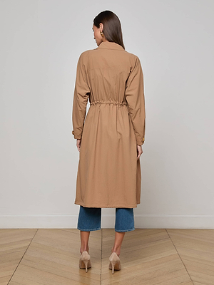 A woman faces away, wearing the L'AGENCE Toshi Windbreaker trench coat with a gathered waist, paired with blue cropped jeans and beige heels, standing on a light wooden floor against a plain white wall.