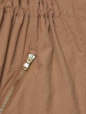 Close-up of the Toshi Windbreaker by L'AGENCE, featuring light brown water-repellent fabric with a slightly crinkled texture and a gold metal zipper sewn diagonally, partially unzipped.