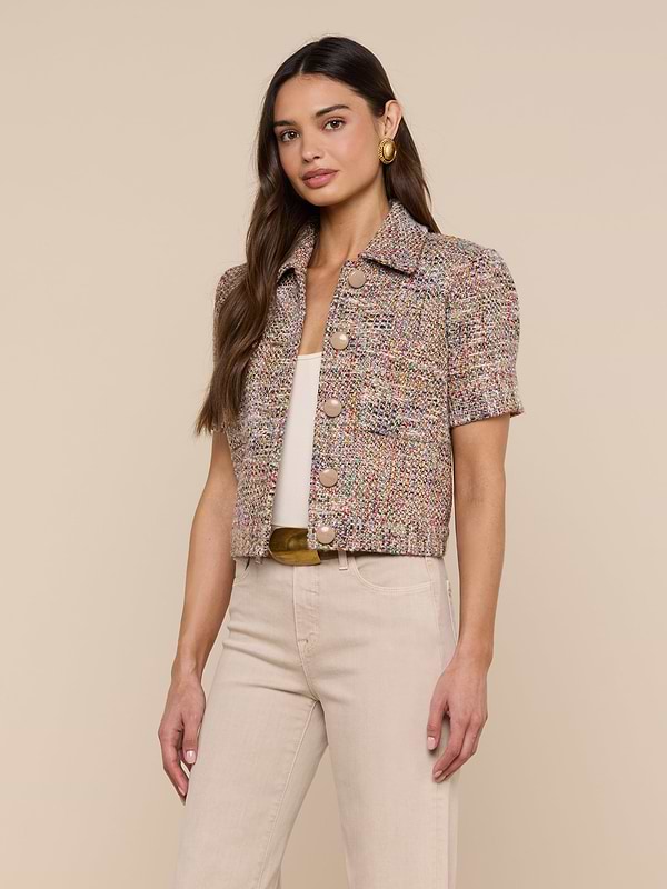 A woman is wearing the Chita Tweed Jacket—a multi-colored, short-sleeved cropped tweed piece—over a white top and beige pants, standing against a plain light background.