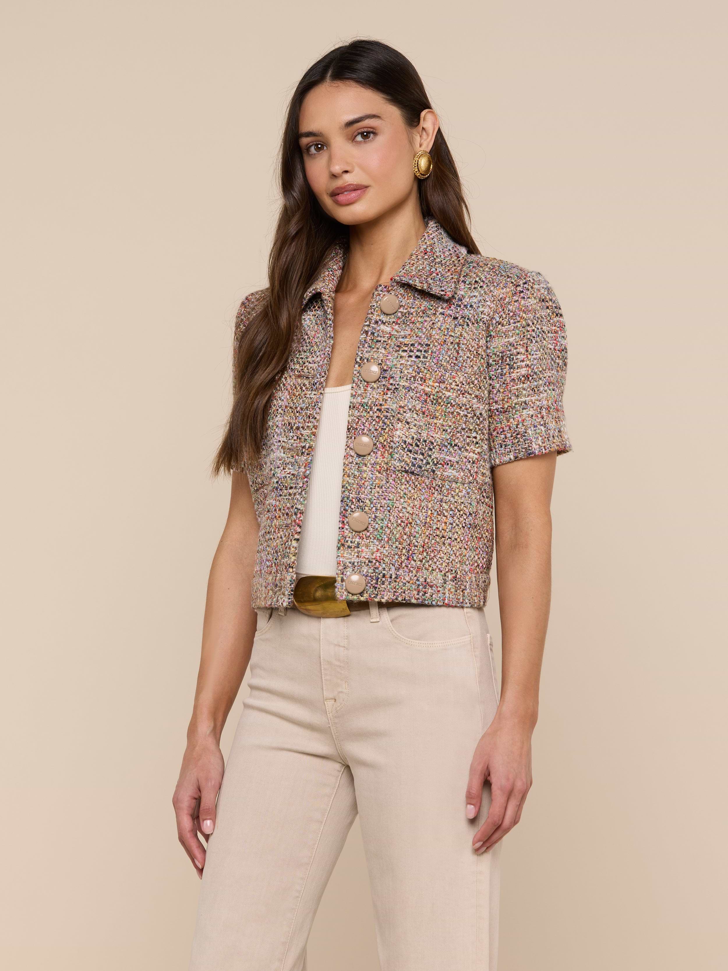 A woman is wearing the Chita Tweed Jacket—a multi-colored, short-sleeved cropped tweed piece—over a white top and beige pants, standing against a plain light background.