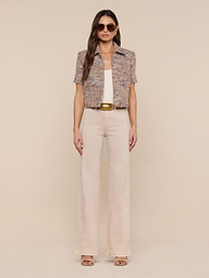 A woman wearing sunglasses, the Chita Tweed Jacket—a short-sleeved multi-colored tweed piece—a white top, beige high-waisted wide-leg pants, and sandals stands against a plain white background.