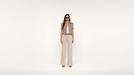 A woman wearing sunglasses, a short-sleeve patterned jacket, a matching top, beige wide-leg pants, and heeled sandals stands against a plain white background.