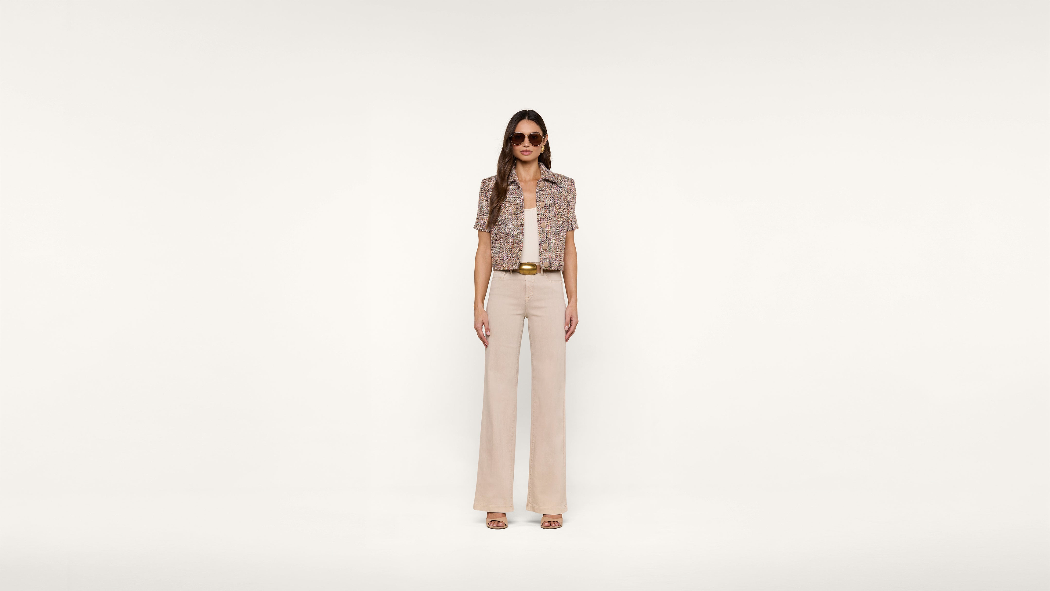 A woman wearing sunglasses, a short-sleeve patterned jacket, a matching top, beige wide-leg pants, and heeled sandals stands against a plain white background.