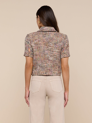 A woman faces away, wearing the Chita Tweed Jacket—a vibrant, multi-colored short-sleeved tweed jacket with a collar—paired with light beige high-waisted pants. Her long brown hair is loose against a plain beige background.