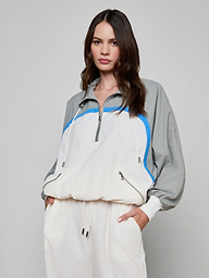 A woman wearing the Aurelia Windbreaker—a light gray and white retro jacket with blue accents—paired with matching white pants, stands with one hand in her pocket against a plain background.