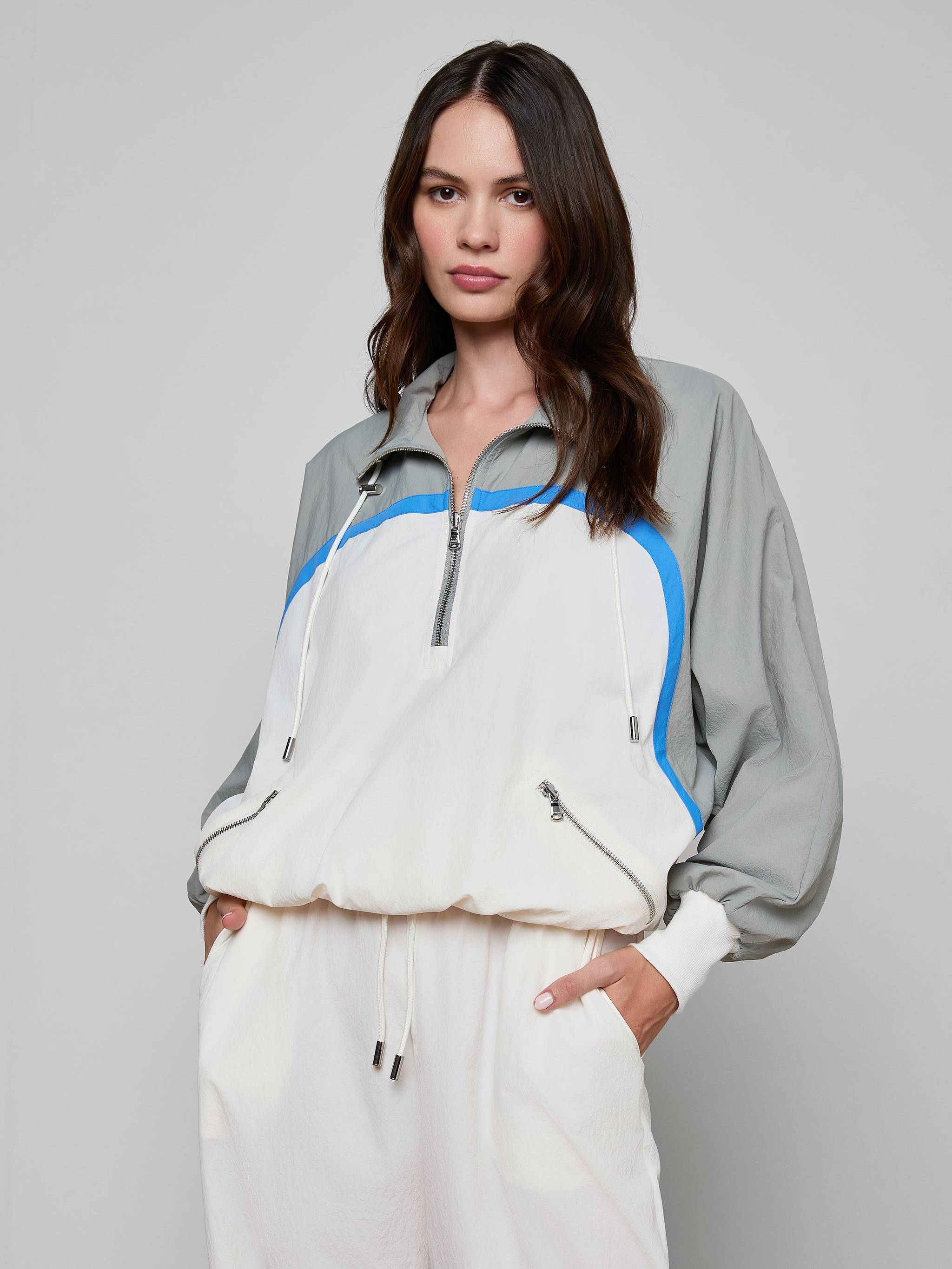 A woman wearing the Aurelia Windbreaker—a light gray and white retro jacket with blue accents—paired with matching white pants, stands with one hand in her pocket against a plain background.