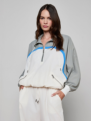 A woman with long brown hair models the Aurelia Windbreaker—a light grey and white zip-up jacket with blue accents—paired with matching white pants, standing hands in pockets against a plain light backdrop.