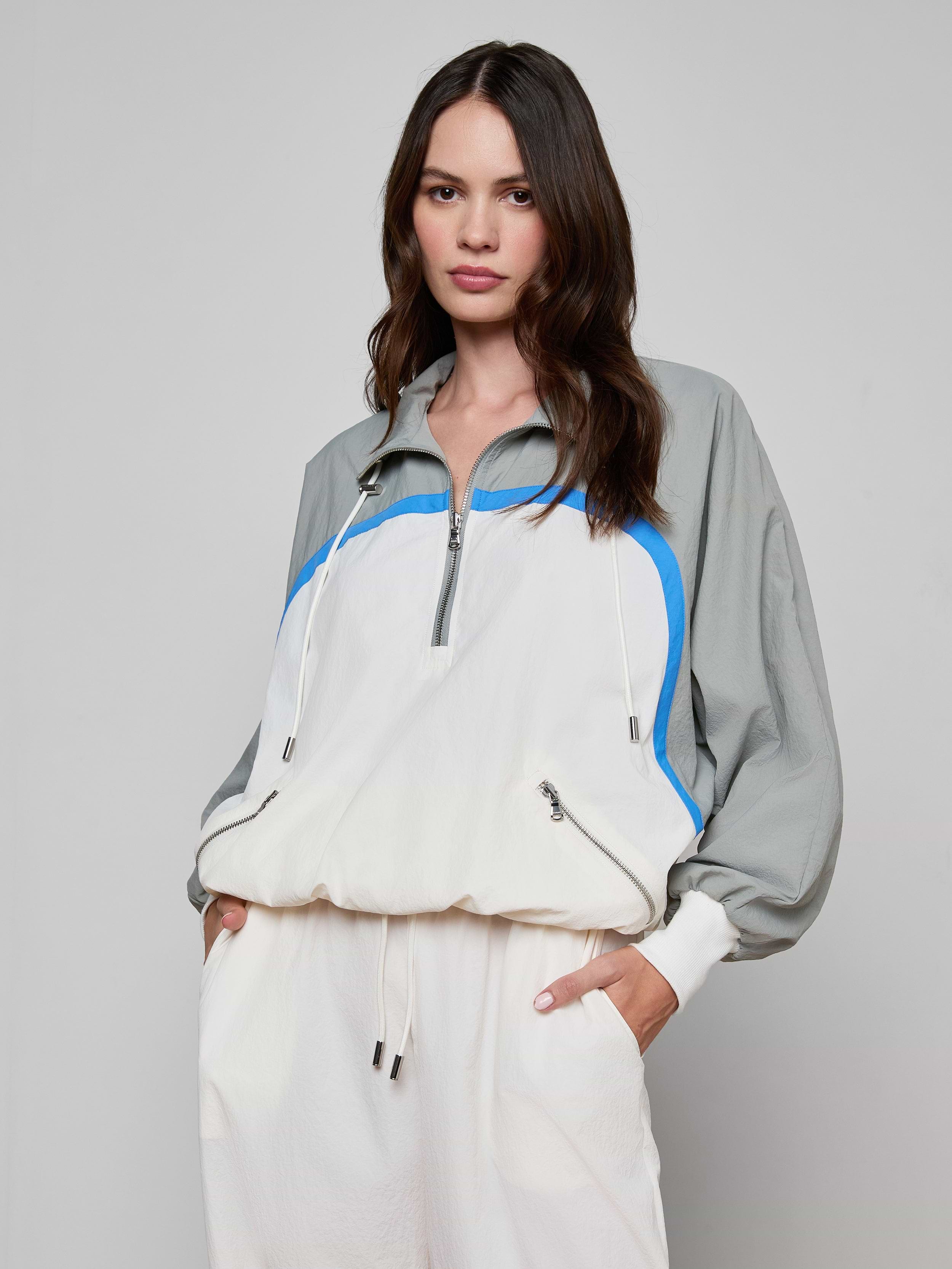 A woman wearing the Aurelia Windbreaker—a light gray and white retro jacket with blue accents—paired with matching white pants, stands with one hand in her pocket against a plain background.