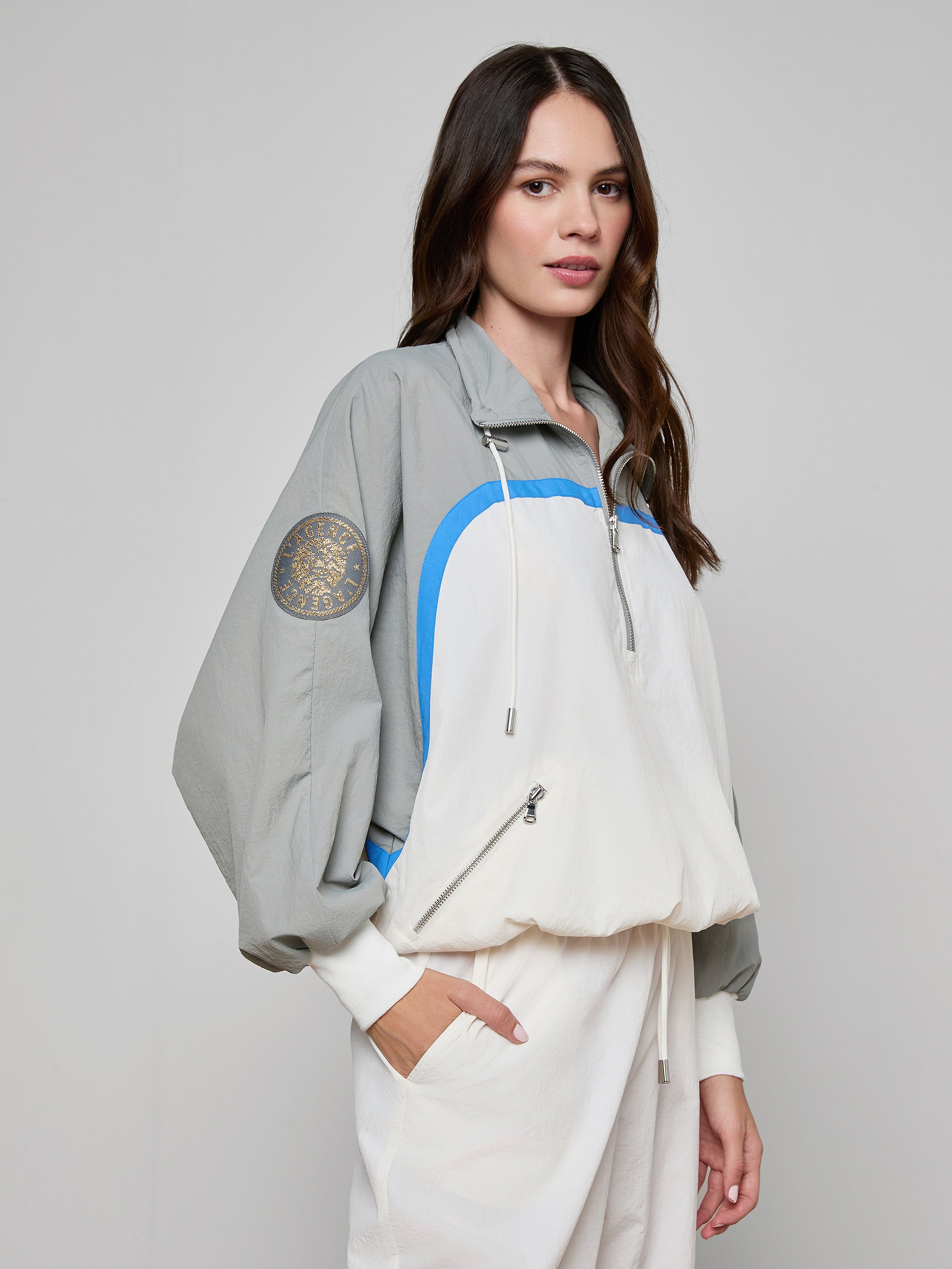 A woman with long brown hair wears the Aurelia Windbreaker—a light gray and white retro jacket with blue accents and an emblem on the sleeve—paired with matching white pants, standing against a plain background.