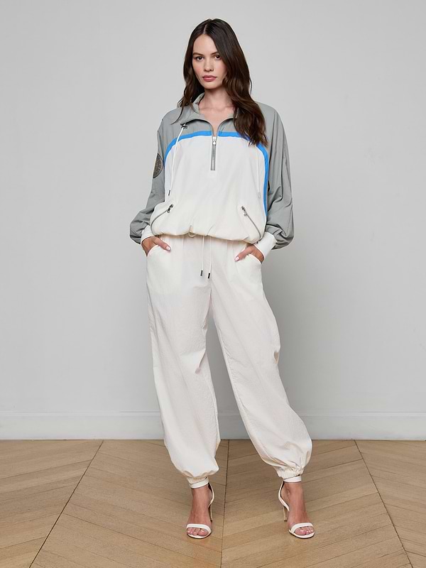 A woman stands facing forward, wearing the Aurelia Windbreaker—a light-colored athleisure tracksuit featuring gray sleeves and blue accents—paired with open-toe heels. The background is plain white.