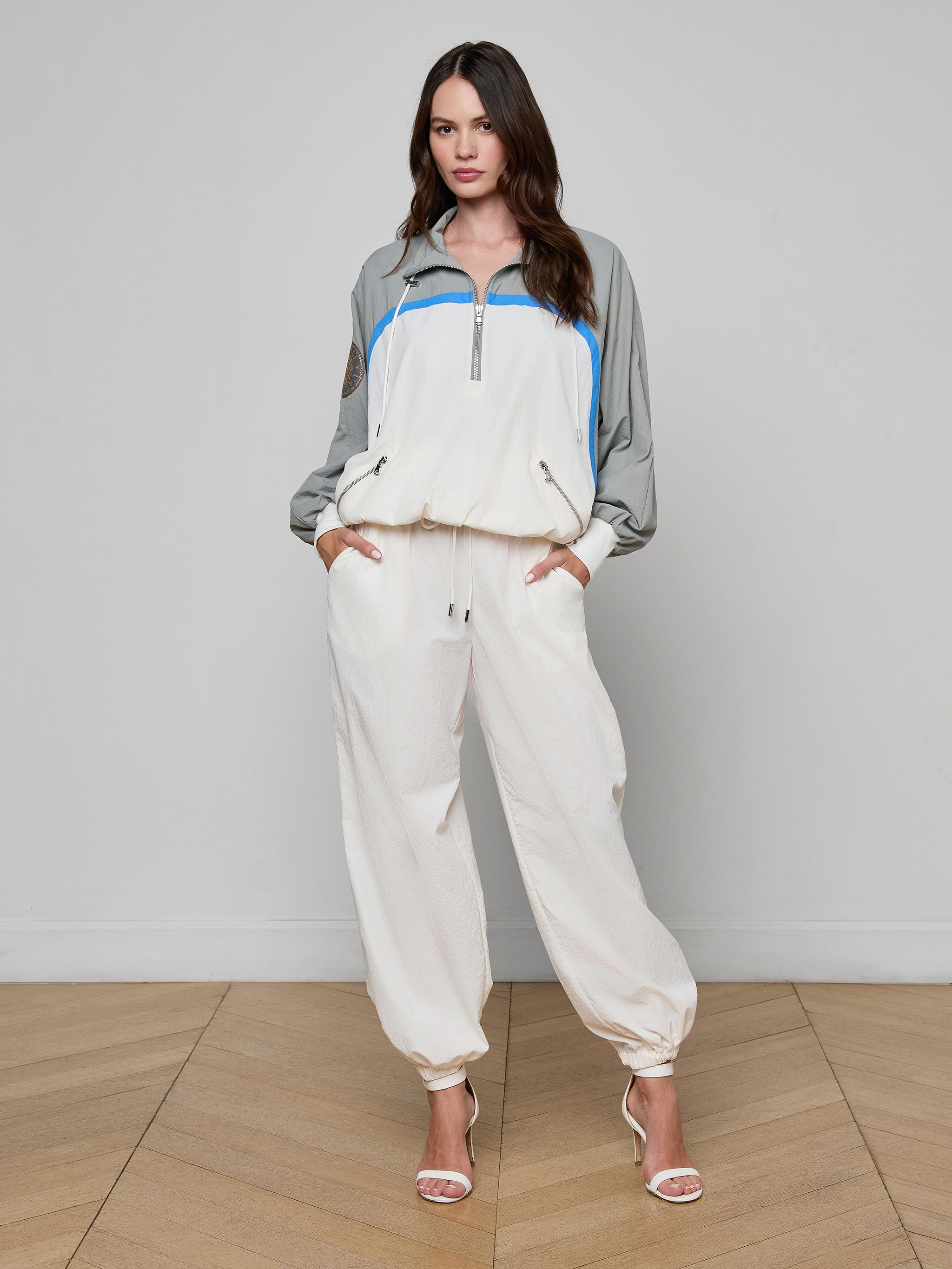 A woman stands facing forward, wearing the Aurelia Windbreaker—a light-colored athleisure tracksuit featuring gray sleeves and blue accents—paired with open-toe heels. The background is plain white.