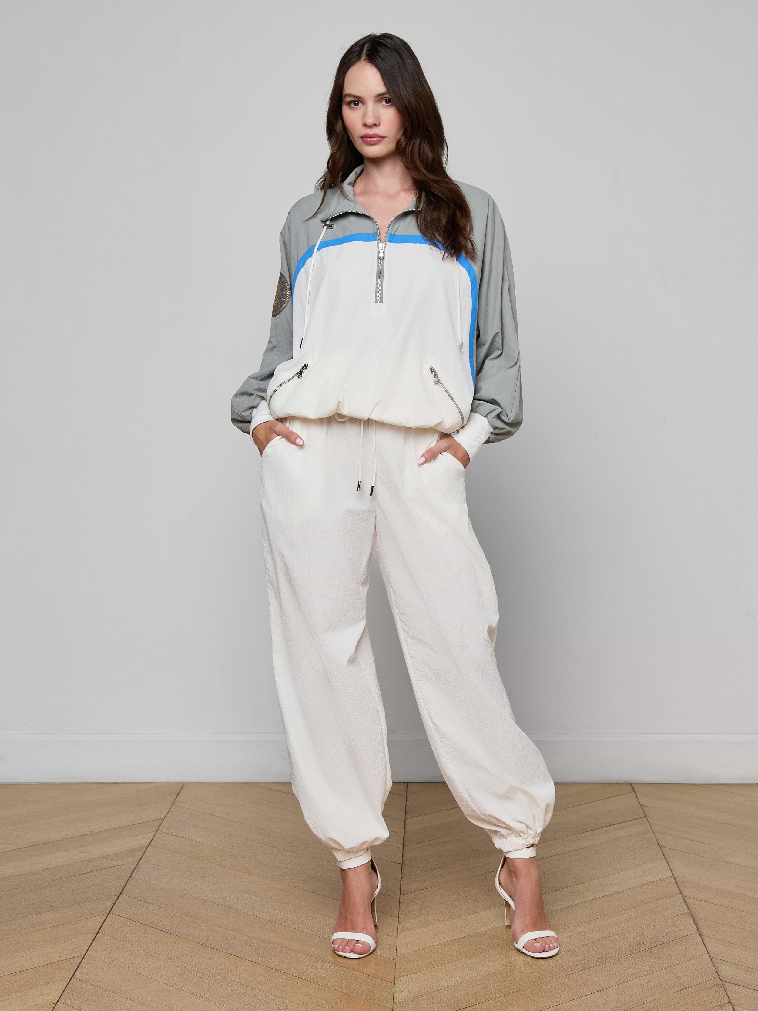 A woman stands facing forward, wearing the Aurelia Windbreaker—a light-colored athleisure tracksuit featuring gray sleeves and blue accents—paired with open-toe heels. The background is plain white.