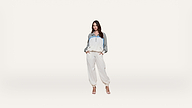 A woman stands against a plain white background wearing a white and gray tracksuit with blue accents and high-heeled sandals, hands in her pockets.