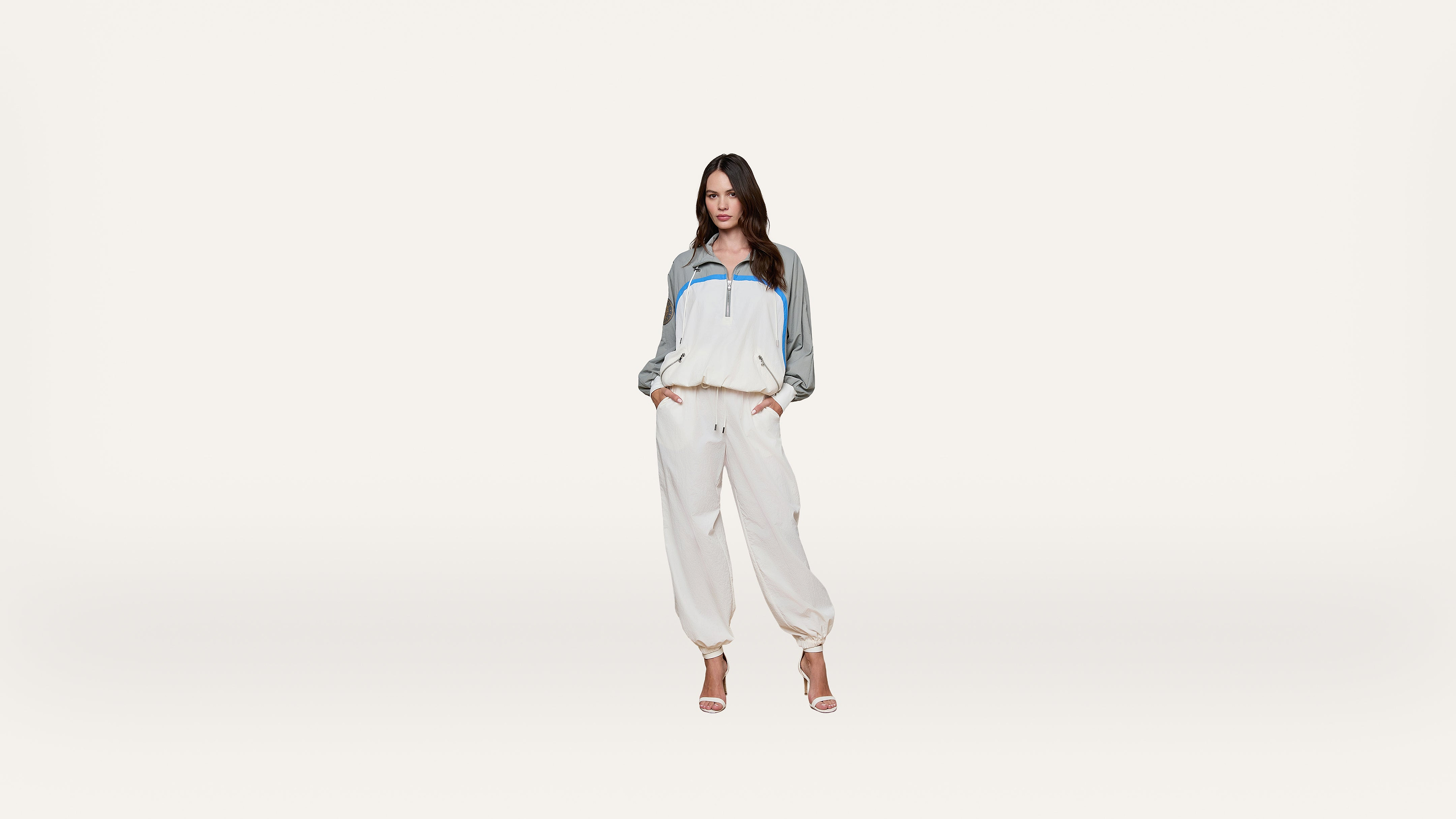 A woman stands against a plain white background wearing a white and gray tracksuit with blue accents and high-heeled sandals, hands in her pockets.
