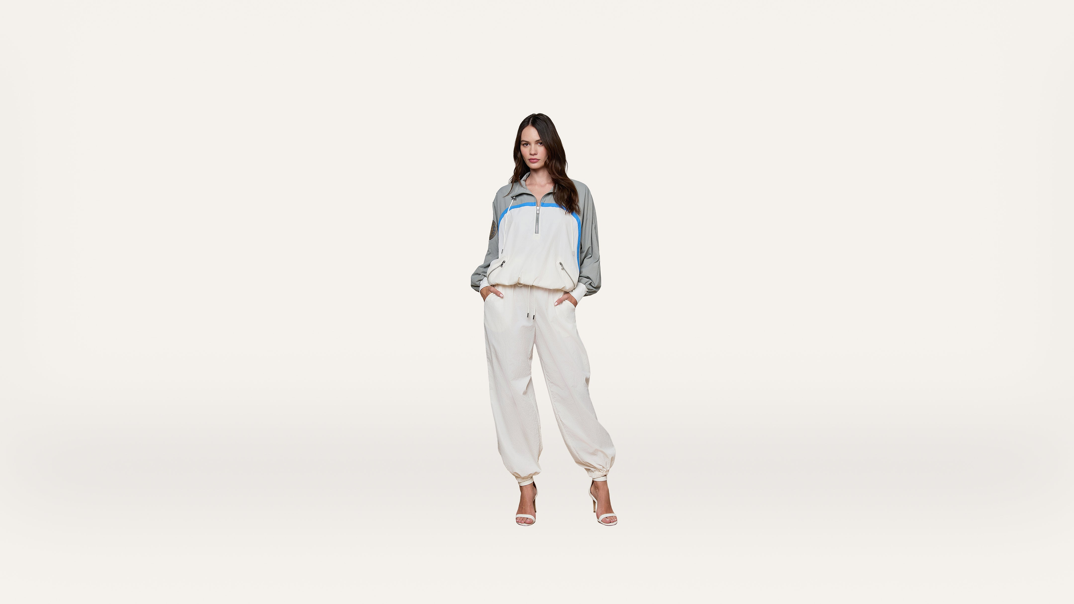 A woman stands against a plain white background wearing a white and gray tracksuit with blue accents and high-heeled sandals, hands in her pockets.