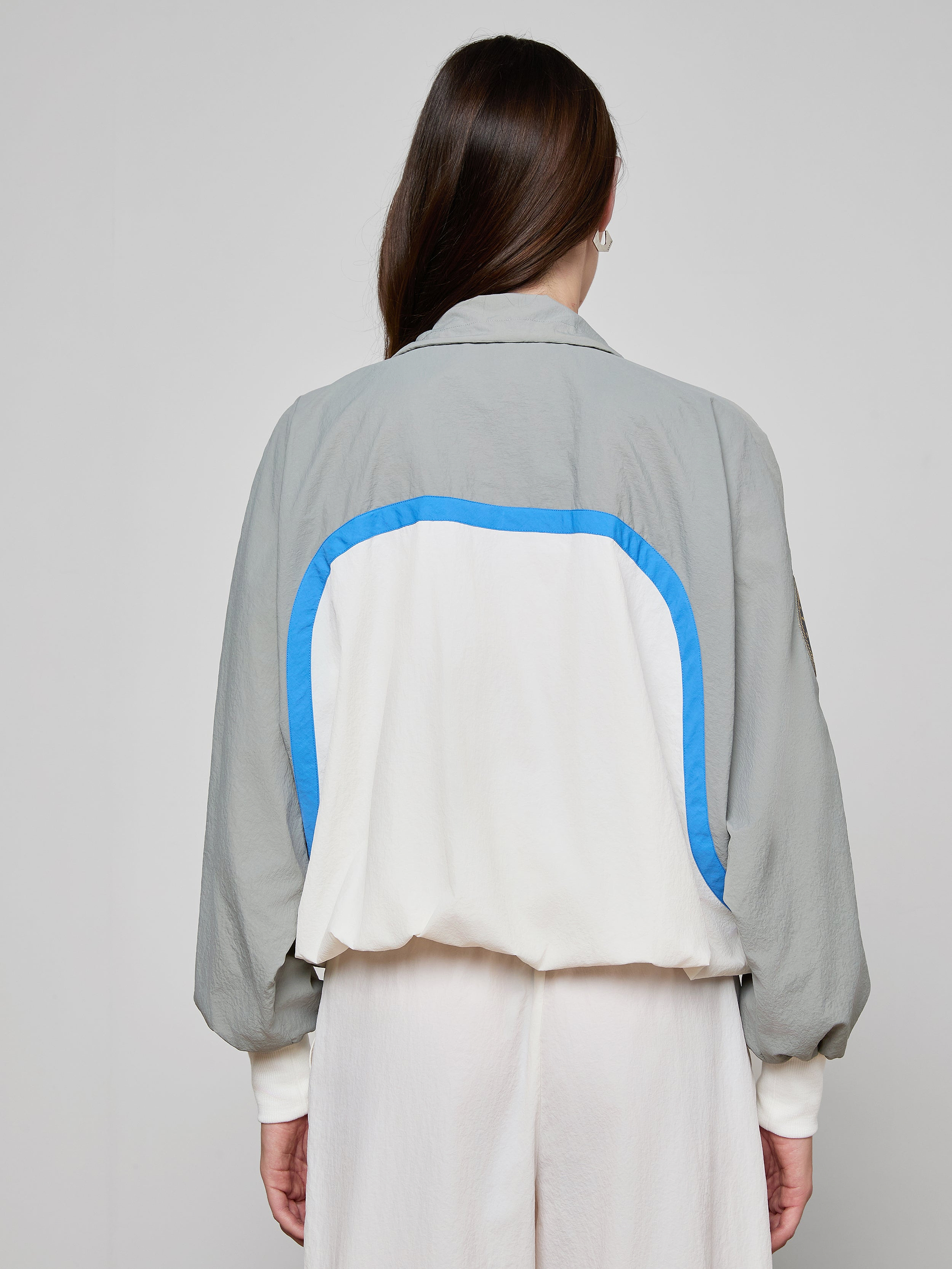 A person with long brown hair, seen from the back, wears the Aurelia Windbreaker—a light gray and white retro jacket featuring a blue curved stripe—paired with loose light-colored pants against a plain background.