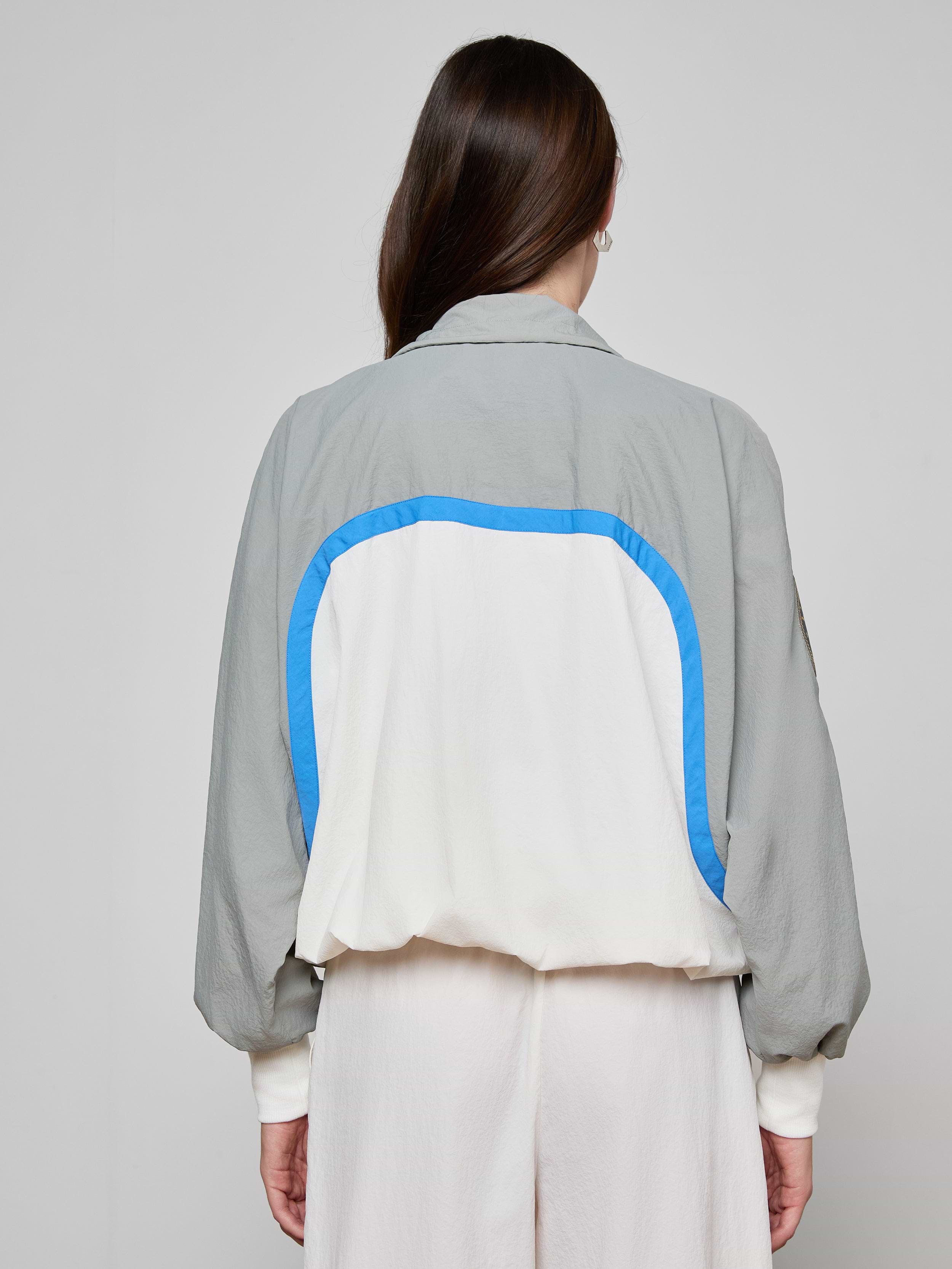 A person with long brown hair, seen from the back, wears the Aurelia Windbreaker—a light gray and white retro jacket featuring a blue curved stripe—paired with loose light-colored pants against a plain background.
