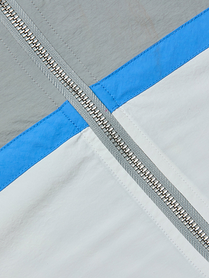 Close-up of the Aurelia Windbreaker’s metallic zipper running diagonally across fabric in light gray, a blue stripe, and white—classic color-blocked details giving this jacket its signature retro-athleisure style.