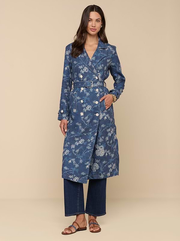 A woman with long brown hair and a soft smile stands against a beige background, wearing the Bradley Linen Trench Coat over wide-leg jeans and brown sandals, highlighting her classic style in this lightweight blue floral-patterned outer layer.