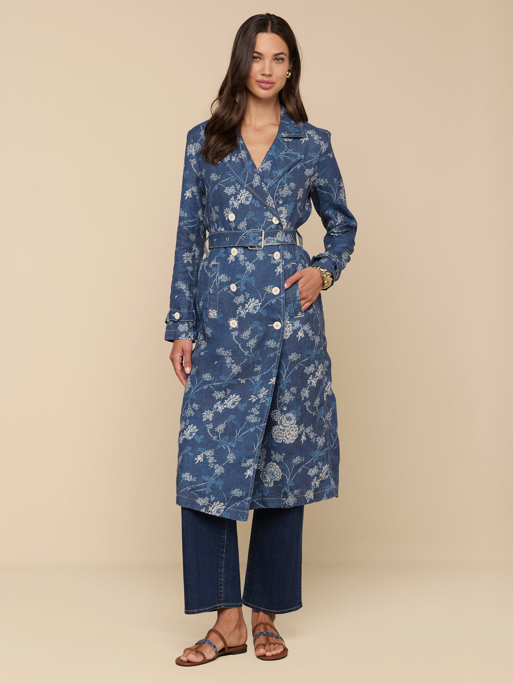 A woman with long brown hair and a soft smile stands against a beige background, wearing the Bradley Linen Trench Coat over wide-leg jeans and brown sandals, highlighting her classic style in this lightweight blue floral-patterned outer layer.