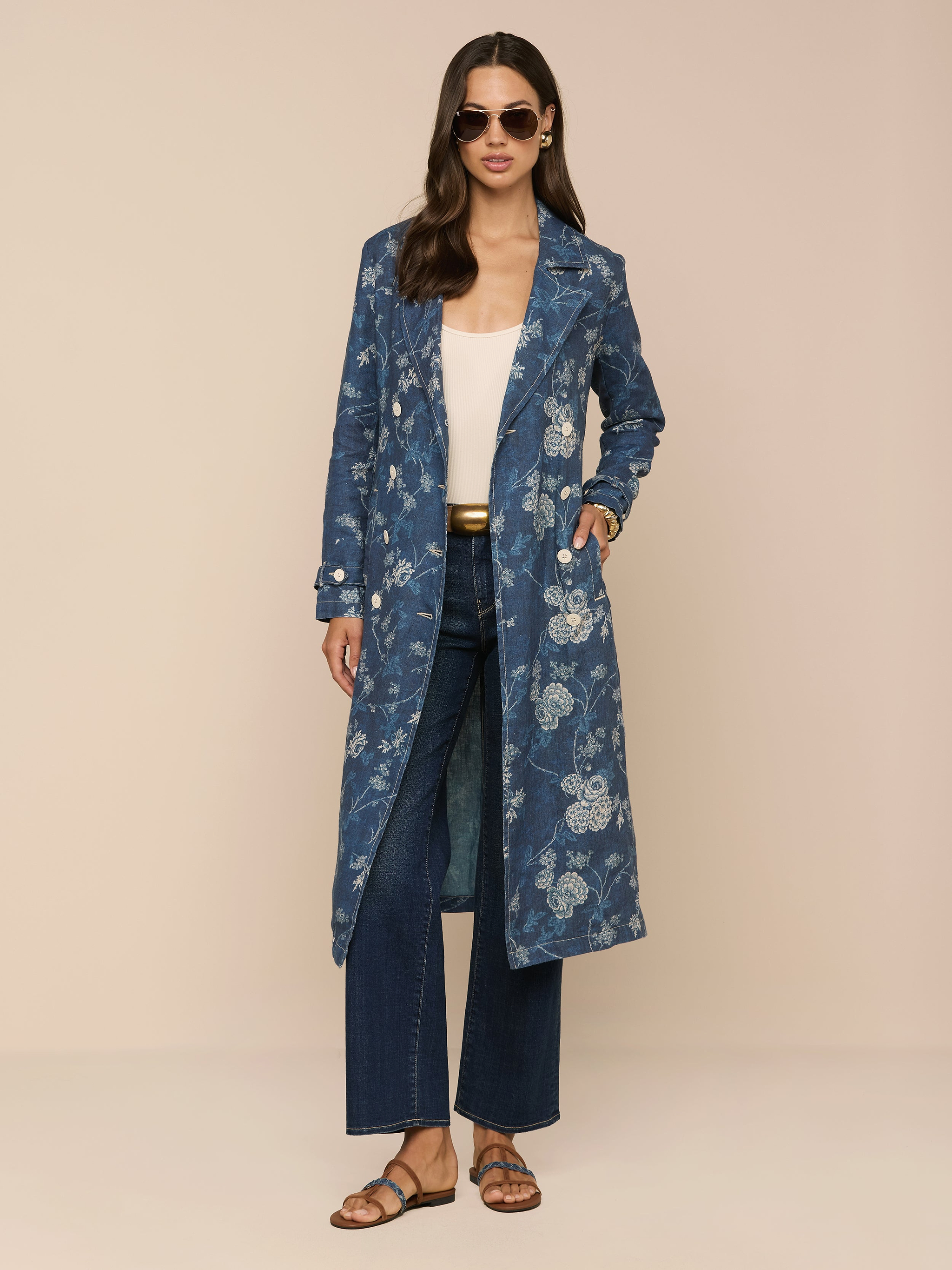 A woman stands against a beige background, showcasing classic style in sunglasses, a white top, wide-leg jeans, brown sandals, and the Bradley Linen Trench Coat—a long blue floral-patterned lightweight outer layer.