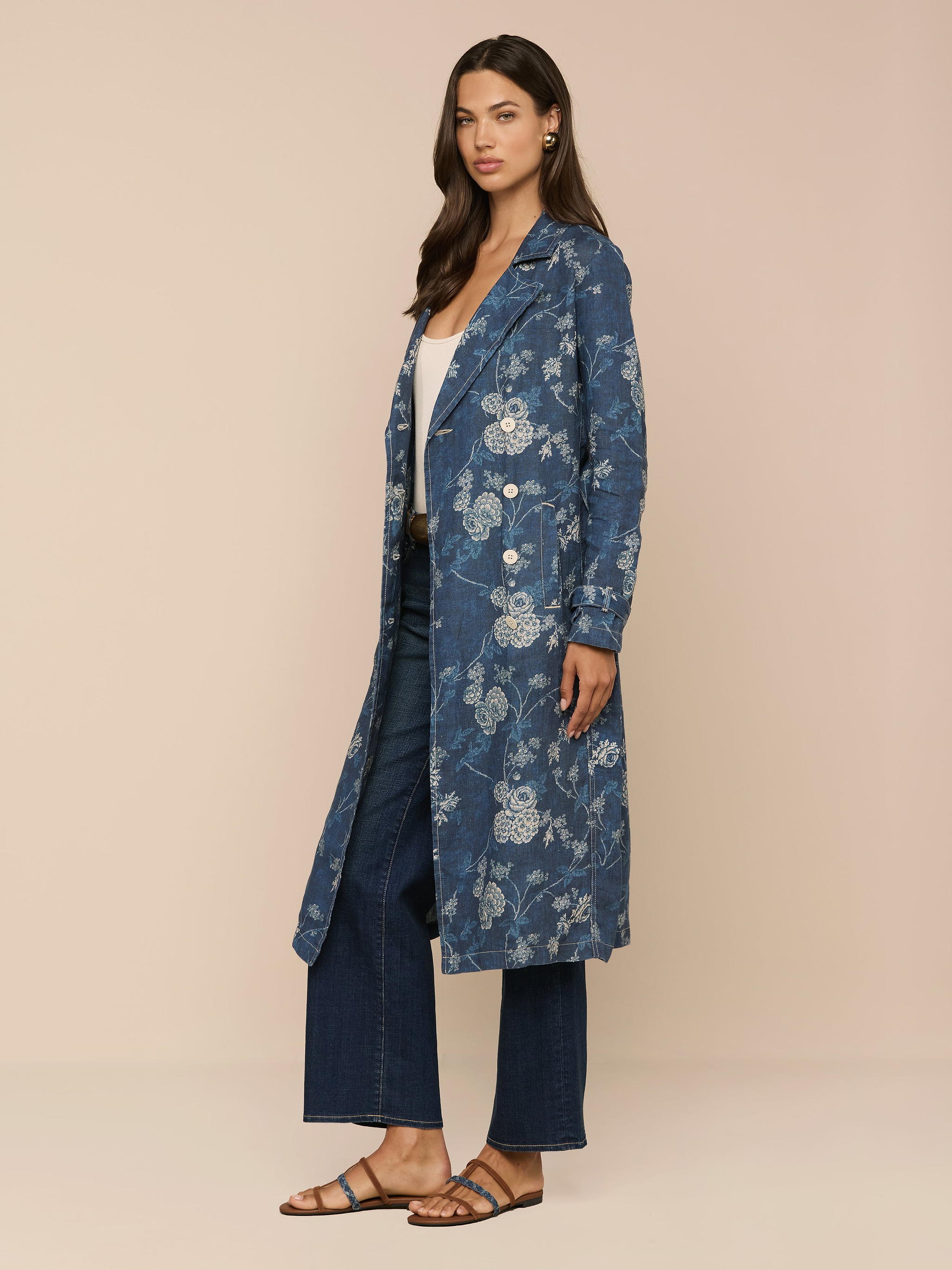 A woman with loose hair and a relaxed expression stands against a beige background, wearing the Bradley Linen Trench Coat in blue floral over a white top, wide-leg dark jeans, and brown sandals.