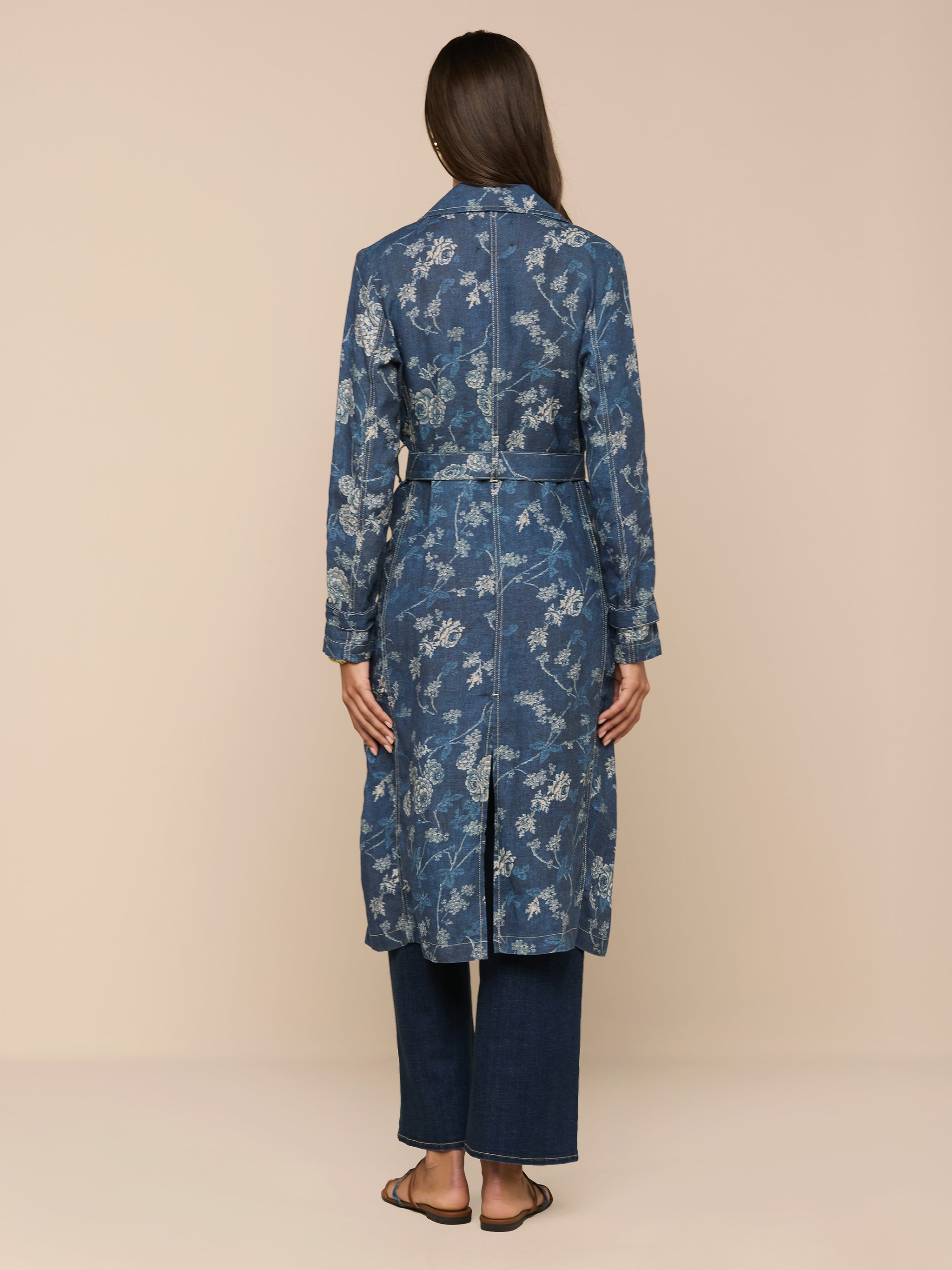 A woman faces away in a classic style, wearing the Bradley Linen Trench Coat—a long blue floral jacket with a matching belt—paired with dark blue pants and sandals against a beige background; an ideal lightweight outer layer.