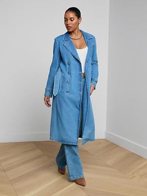 A woman stands indoors wearing the Bradley Trench Coat in medium wash denim over a white top and matching denim pants, paired with brown heeled shoes. She looks to the side with one hand relaxed by her side.