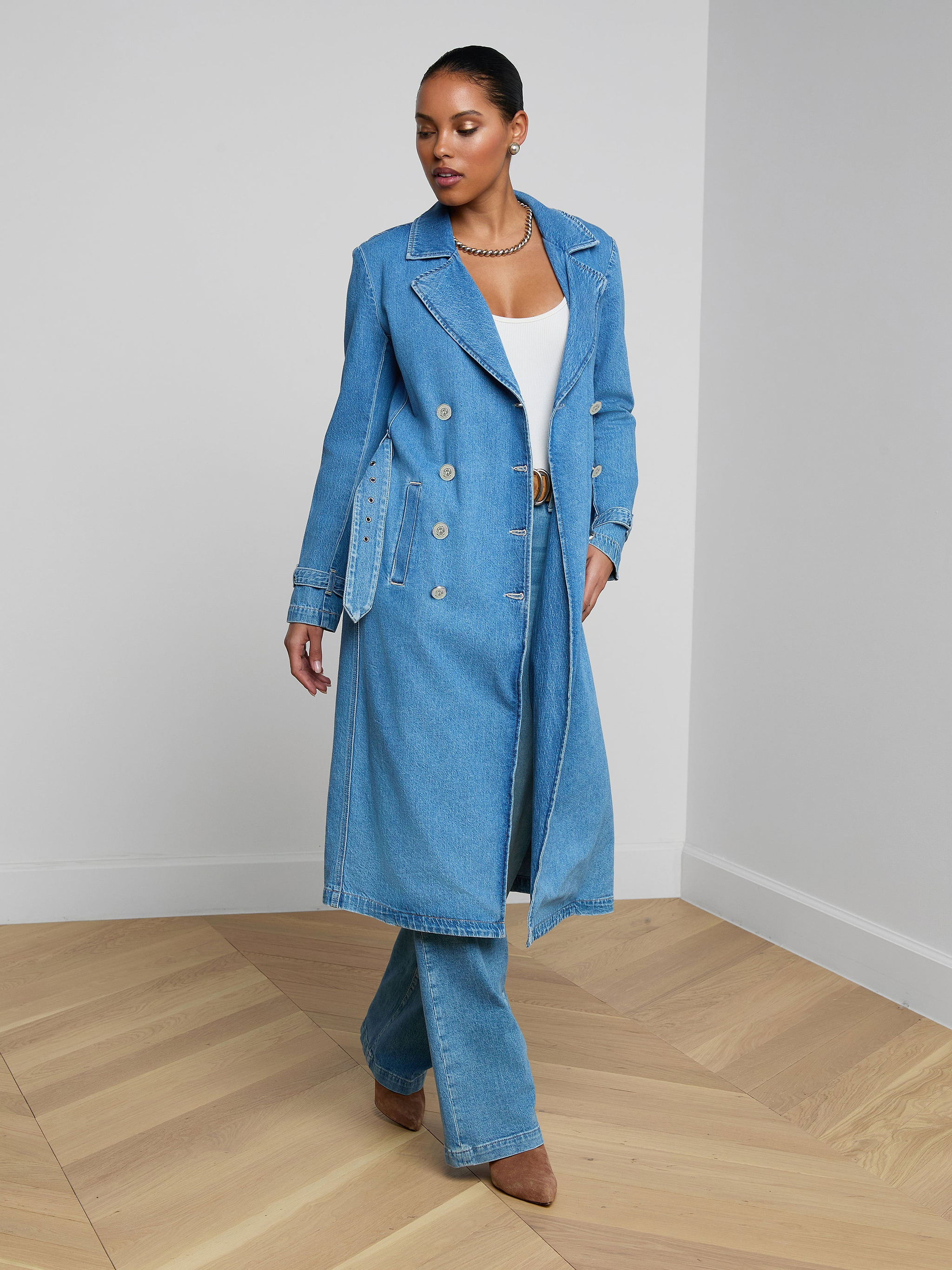 Wearing the Bradley Trench Coat in a lightweight, medium-wash denim, a woman stands against a plain white background, styled with matching denim pants and a white top.