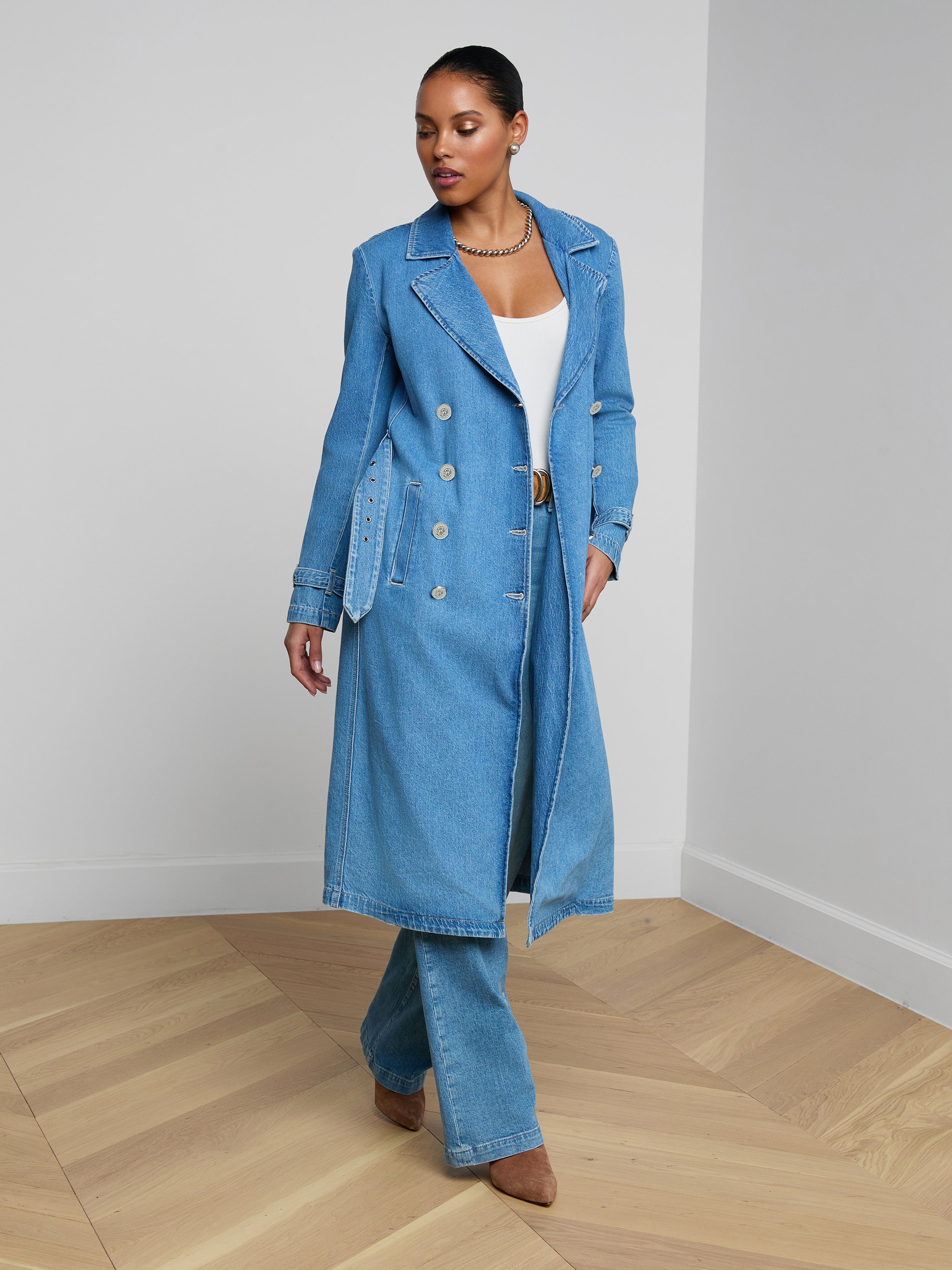 Wearing the Bradley Trench Coat in a lightweight, medium-wash denim, a woman stands against a plain white background, styled with matching denim pants and a white top.