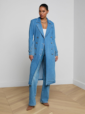 A woman stands in a minimalist room, wearing the Bradley Trench Coat—a medium wash, double-breasted denim trench—over a white top and matching denim pants with tan heels. She gazes forward, hands resting on her coat.