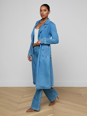 A woman models the Bradley Trench Coat—a lightweight denim style—paired with matching medium-wash jeans, a white top, and tan heels, standing on a wooden floor against a plain white wall.