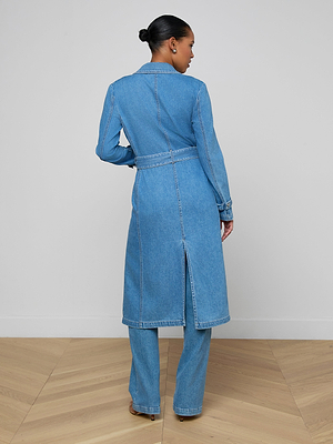 A woman stands with her back to the camera, wearing the Bradley Trench Coat—a medium wash denim trench with a belt—over matching pants. Her hair is in a neat bun as she stands on a wooden floor against a plain light wall.