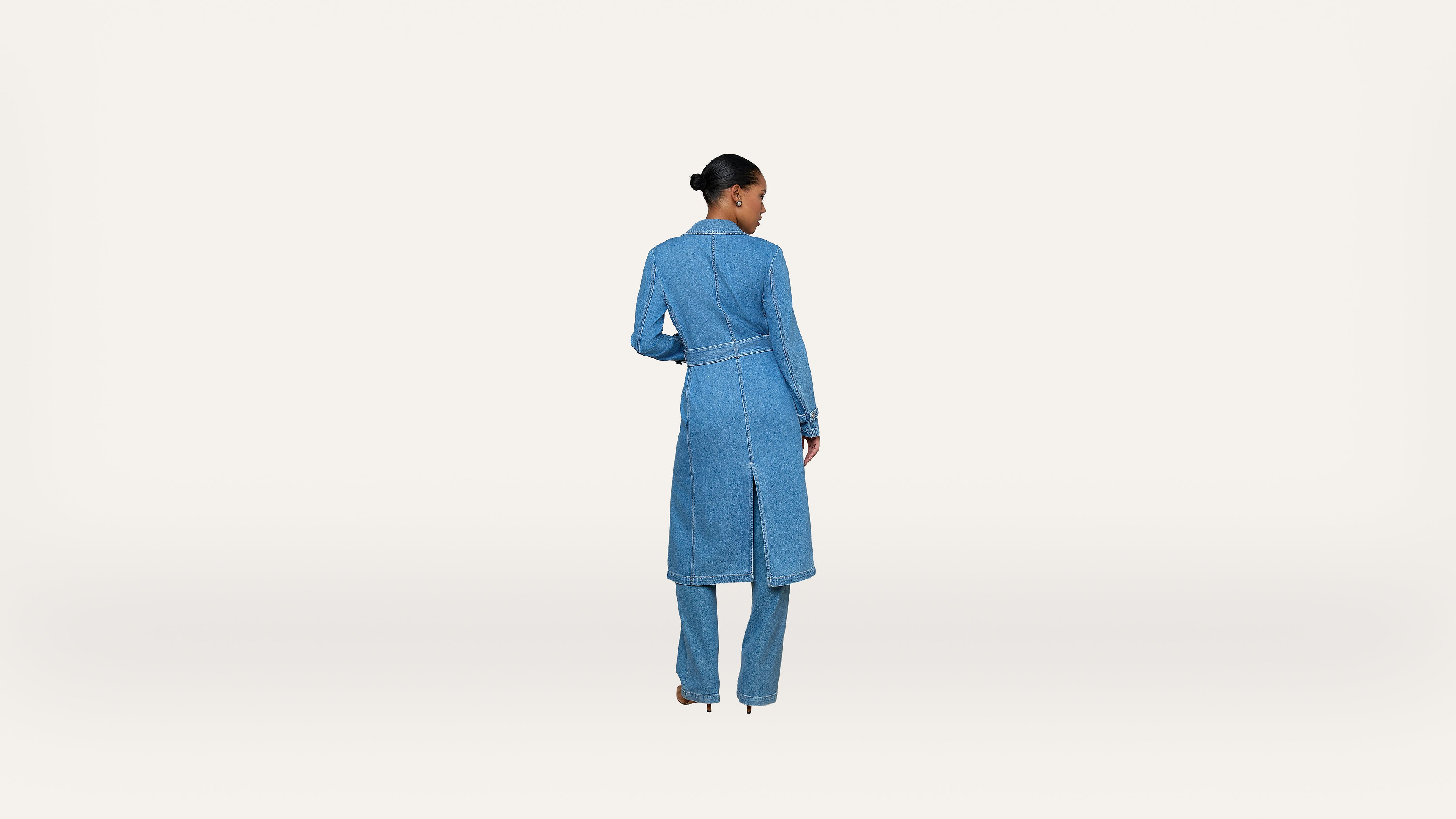 A person stands facing away, wearing a long-sleeved blue denim coat with matching blue pants and heels. Their hair is pulled back neatly, and the background is plain white.
