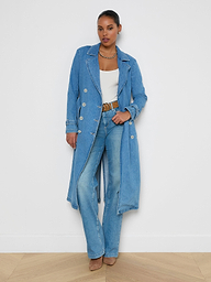 A woman wearing the Bradley Trench Coat—a long, medium wash denim trench—paired with a white top, light blue jeans, brown belt and shoes, and layered necklaces stands confidently against a plain white background.