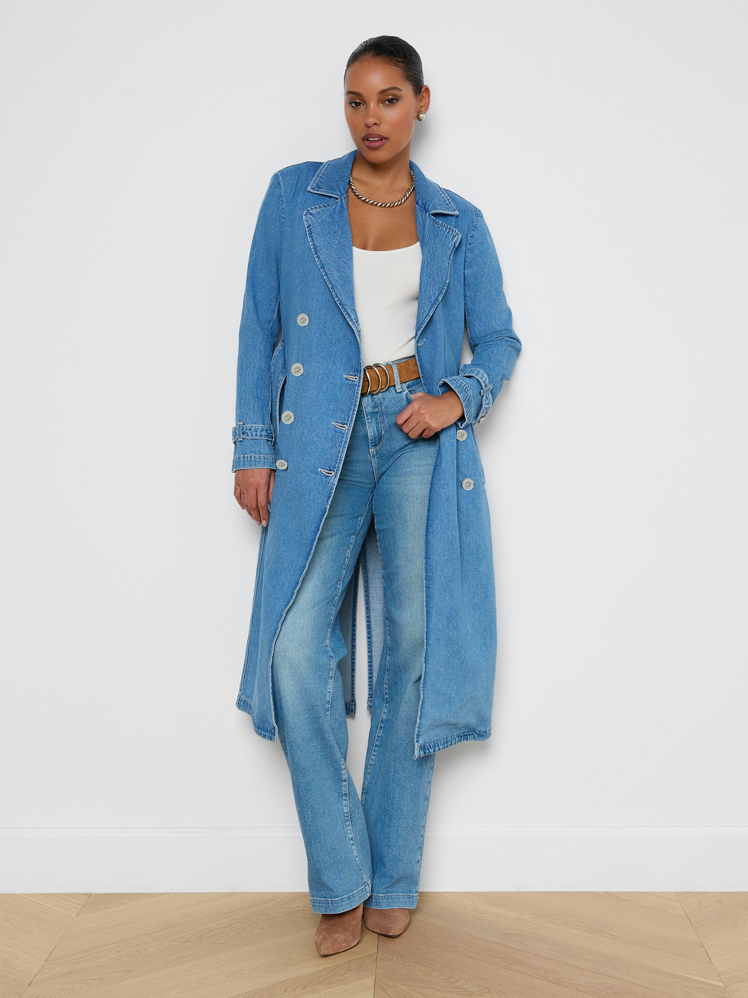 A woman wearing the Bradley Trench Coat—a long, medium wash denim trench—paired with a white top, light blue jeans, brown belt and shoes, and layered necklaces stands confidently against a plain white background.