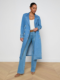 A woman stands wearing the Bradley Trench Coat in a lightweight, medium wash denim over a white top and blue jeans with brown shoes, looking down and gently holding her coat open.