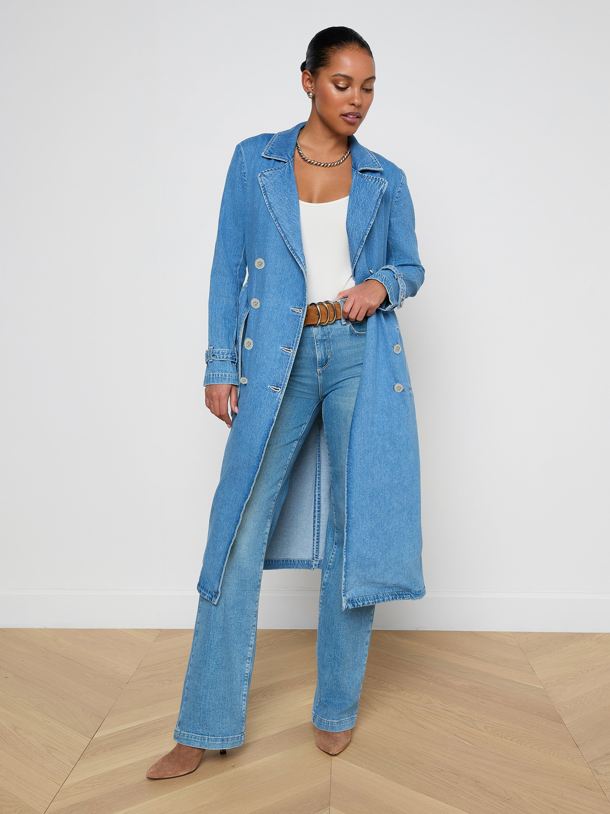 A woman stands wearing the Bradley Trench Coat in a lightweight, medium wash denim over a white top and blue jeans with brown shoes, looking down and gently holding her coat open.