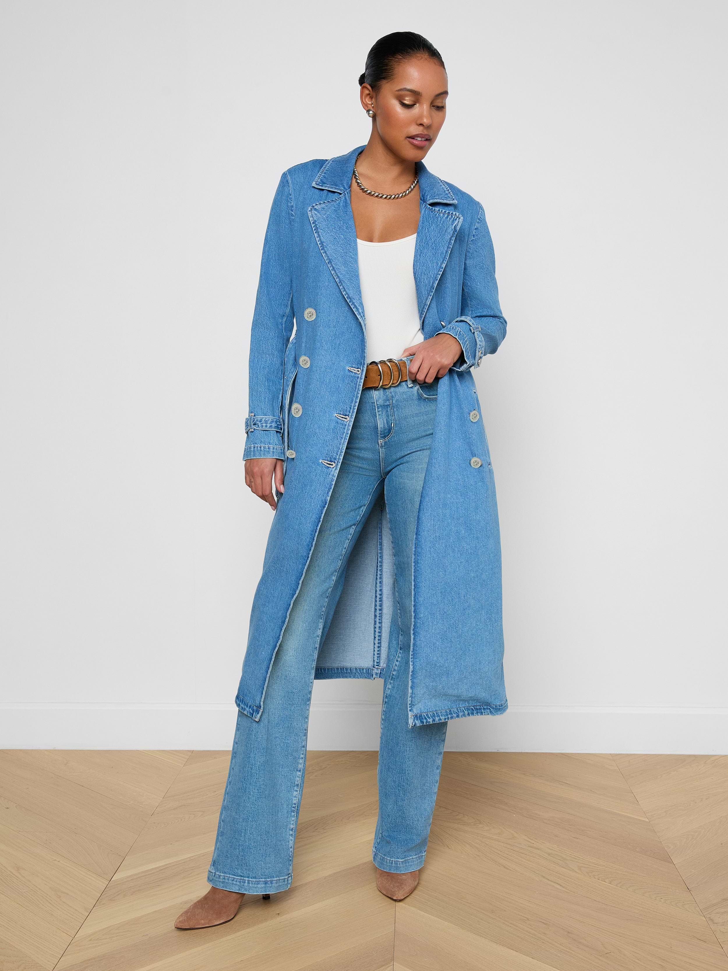 A woman stands wearing the Bradley Trench Coat in a lightweight, medium wash denim over a white top and blue jeans with brown shoes, looking down and gently holding her coat open.