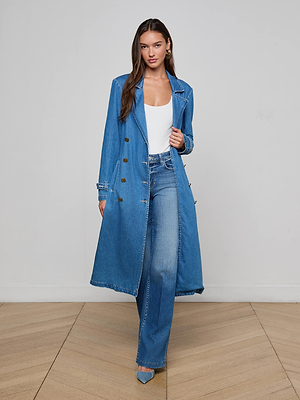 A woman with long brown hair poses indoors against a plain background, wearing the Bradley Trench Coat over a white top and high-waisted blue jeans, paired with blue pointed-toe heels.