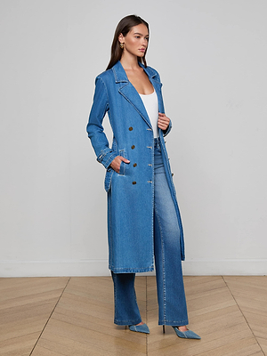 A woman stands on a wooden floor in the Bradley Trench Coat, matching flared jeans, a white top, and blue pointed heels, with her hand in her pocket against a plain light background.