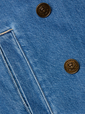 Close-up of the Bradley Trench Coat in light wash blue denim, highlighting a slanted pocket and two brass eagle-embossed buttons for stylish detail on this lightweight design.