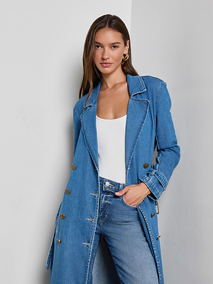 A woman with long brown hair wears the Bradley Trench Coat in light wash denim over a white top and blue jeans, standing against a plain light gray wall with one hand in her pocket, looking at the camera.